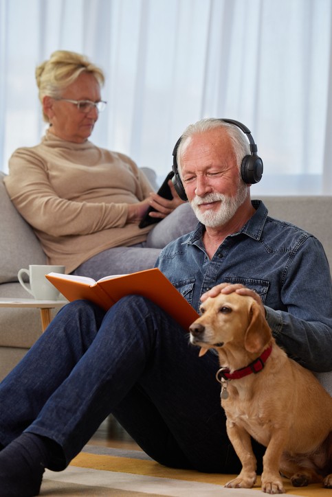 Elderly couple spending leisure time at home with their dog. Spouses reading book, listening to music, podcast or watching a movie while sitting on sofa. Happy retirement concept.